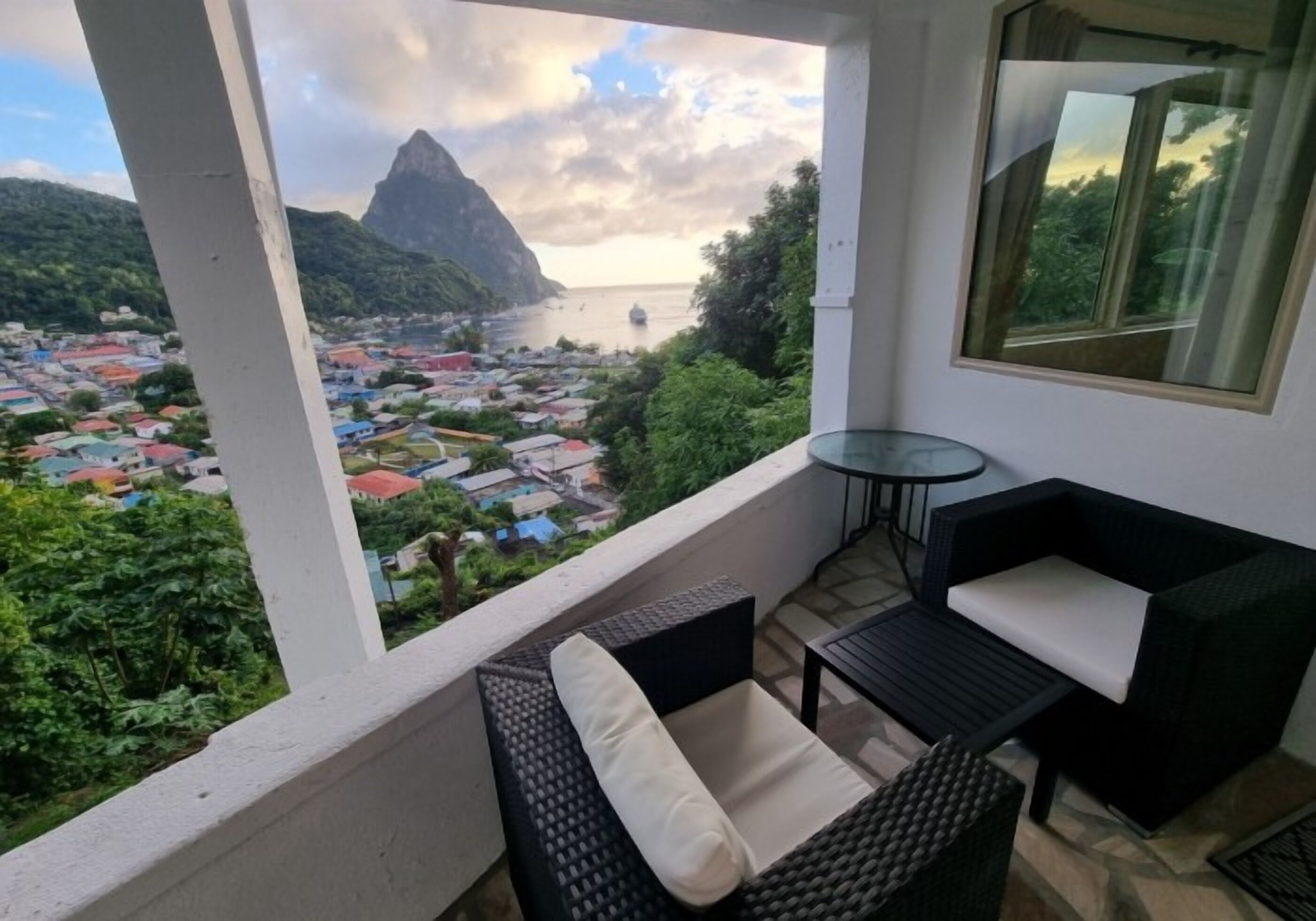 Un balcon agrémenté de meubles en osier, d’une table en verre et offrant une vue sur une ville côtière et une chaîne de montagnes.