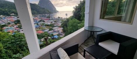 Un balcon agrémenté de meubles en osier, d’une table en verre et offrant une vue sur une ville côtière et une chaîne de montagnes.