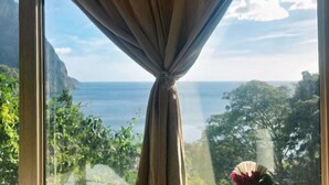 A window with a curtain tied at the top, overlooking a coastal landscape with greenery and a view of the sea. - Enchanting Soufrière Stay | Spa, Pool & Breakfast + Breathtaking Pitons Hikes (Palmiste)