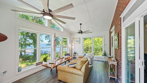 Ein geräumiges Wohnzimmer mit einer großen Couch, einem Holztisch und einem Deckenventilator. Große Fenster bieten einen Blick ins Freie.