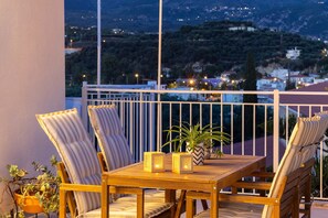 A balcony with a wooden table and striped chairs, a potted plant, and a view of a town at dusk. - Kalamata Cozy Nest With Panoramic Views (Kalamata)