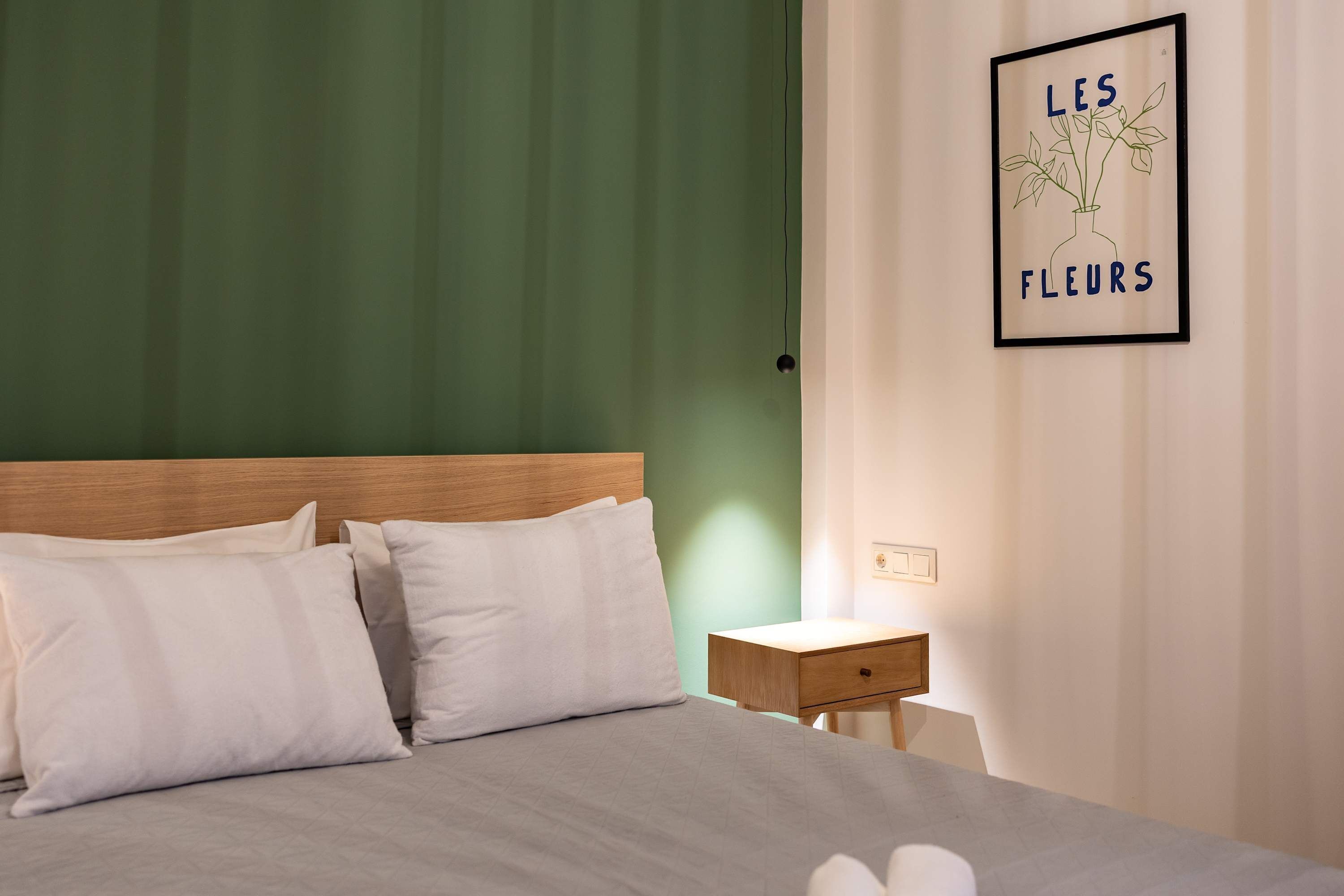 A neatly made bed with white linens, a wooden headboard, and a small bedside table with a lamp. A framed picture of flowers hangs on the wall.