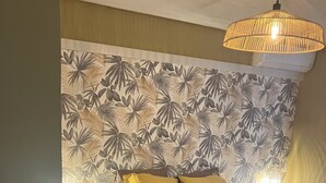 Une chambre à coucher avec un lit, des oreillers jaunes, une table de chevet avec une lampe et un mur orné d’un motif de feuilles tropicales.