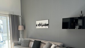 A modern living room with a beige sectional sofa, a flat-screen TV mounted on the wall, and a window with curtains.