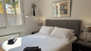 A neatly made bed with white linens and two pillows, a bedside lamp, a small table with a lamp, a framed artwork on the wall, and a window with a view of buildings.