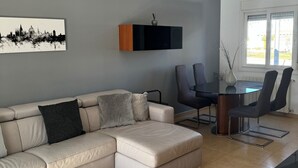 A modern living room with a sectional sofa, a dining table with chairs, a ceiling fan, and a cityscape artwork on the wall.
