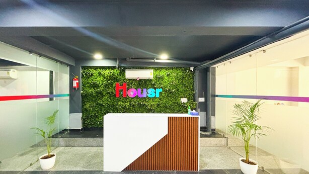 Un comptoir d’accueil avec un mur vert et le mot « House » écrit en lettres colorées. Deux plantes en pot sont placées de chaque côté du comptoir.