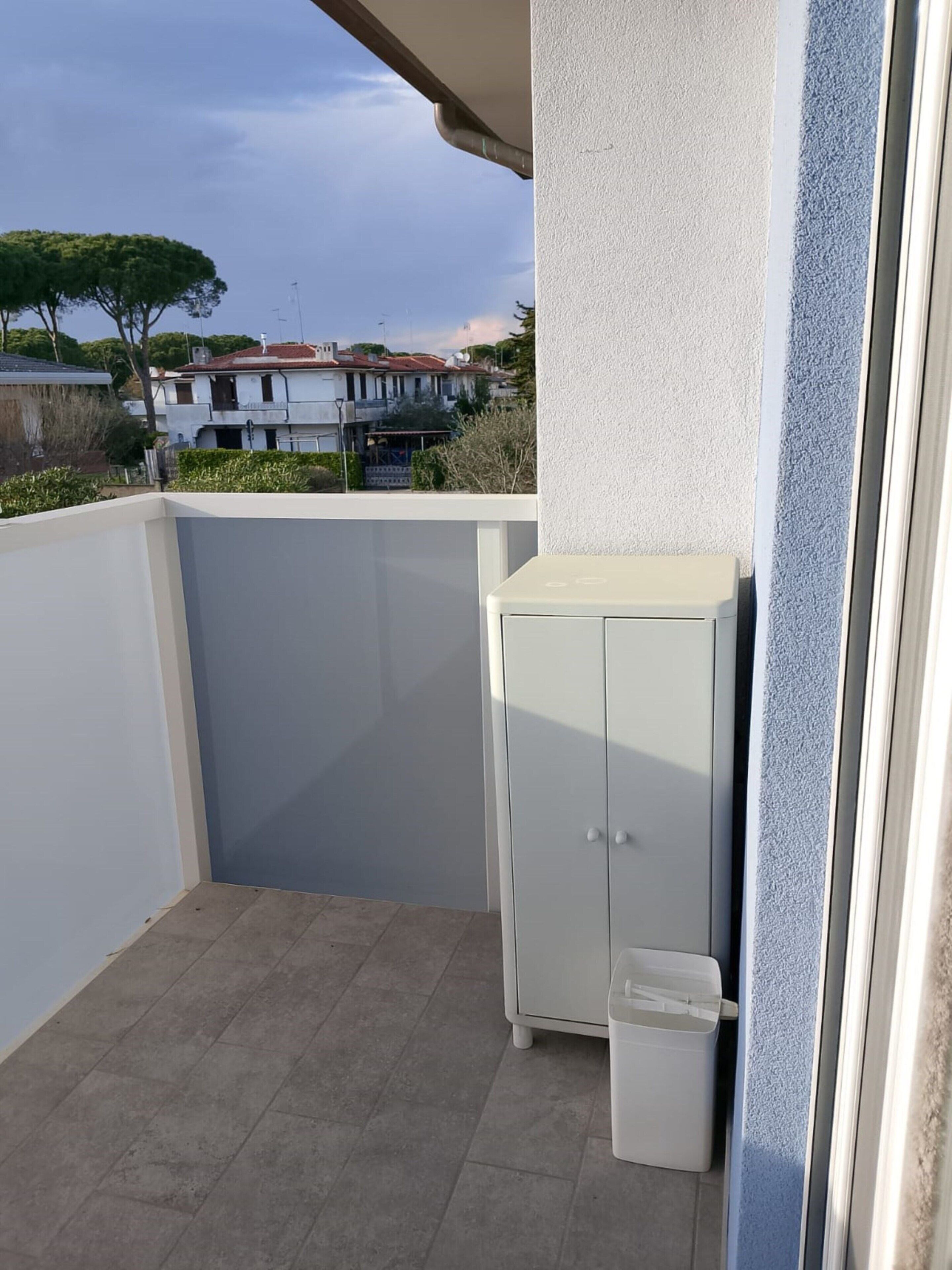 A balcony with a white cabinet and a trash bin, a tiled floor, and a view of houses in the background.