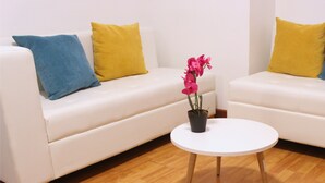 Una habitación con dos sofás blancos, cada uno con un cojín azul y amarillo, y una pequeña mesa blanca con una planta en maceta en el centro.