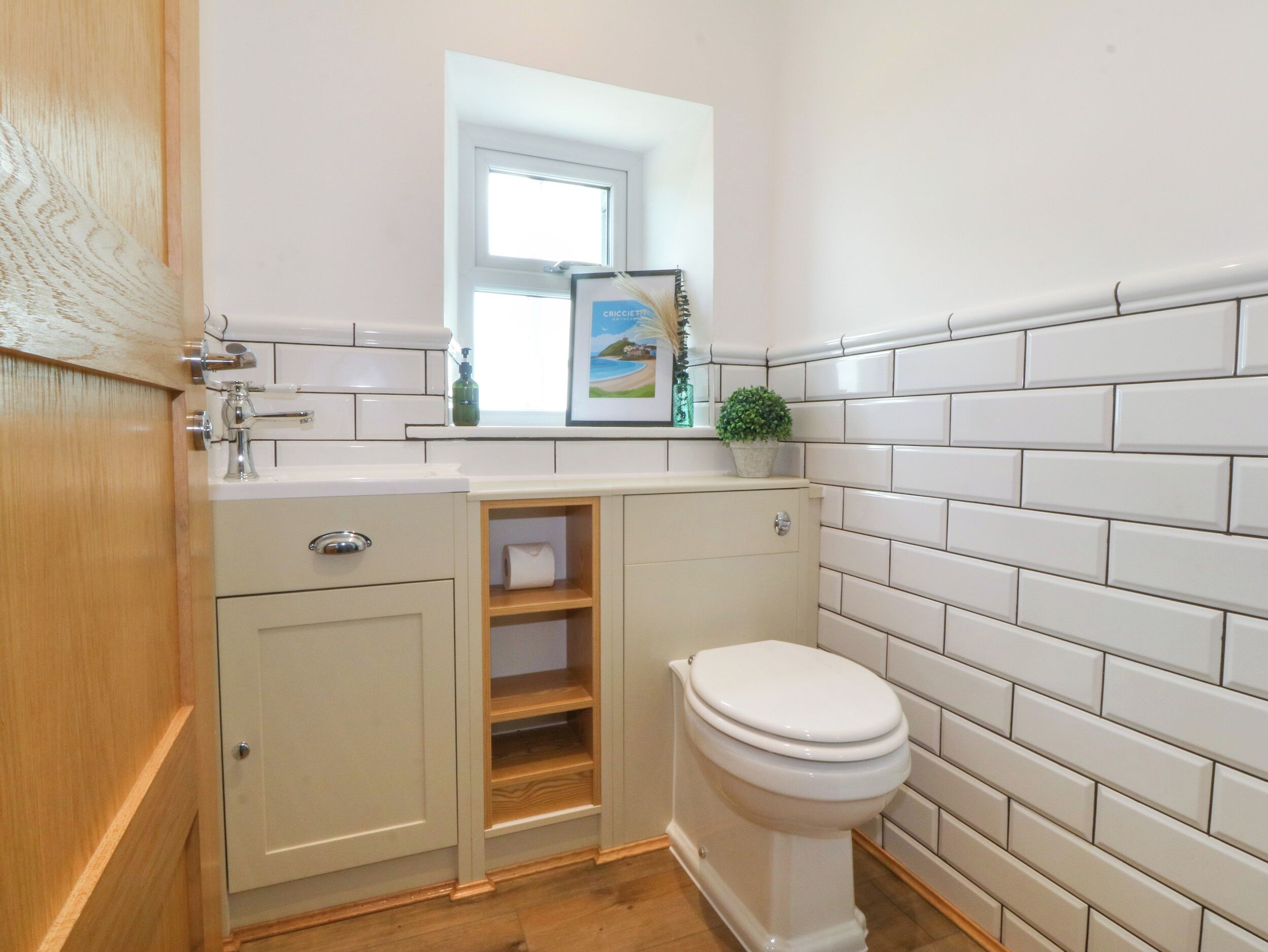Une salle de bain comprenant des toilettes, un lavabo avec un miroir au-dessus, et une petite étagère ornée d’une plante et d’un tableau encadré.