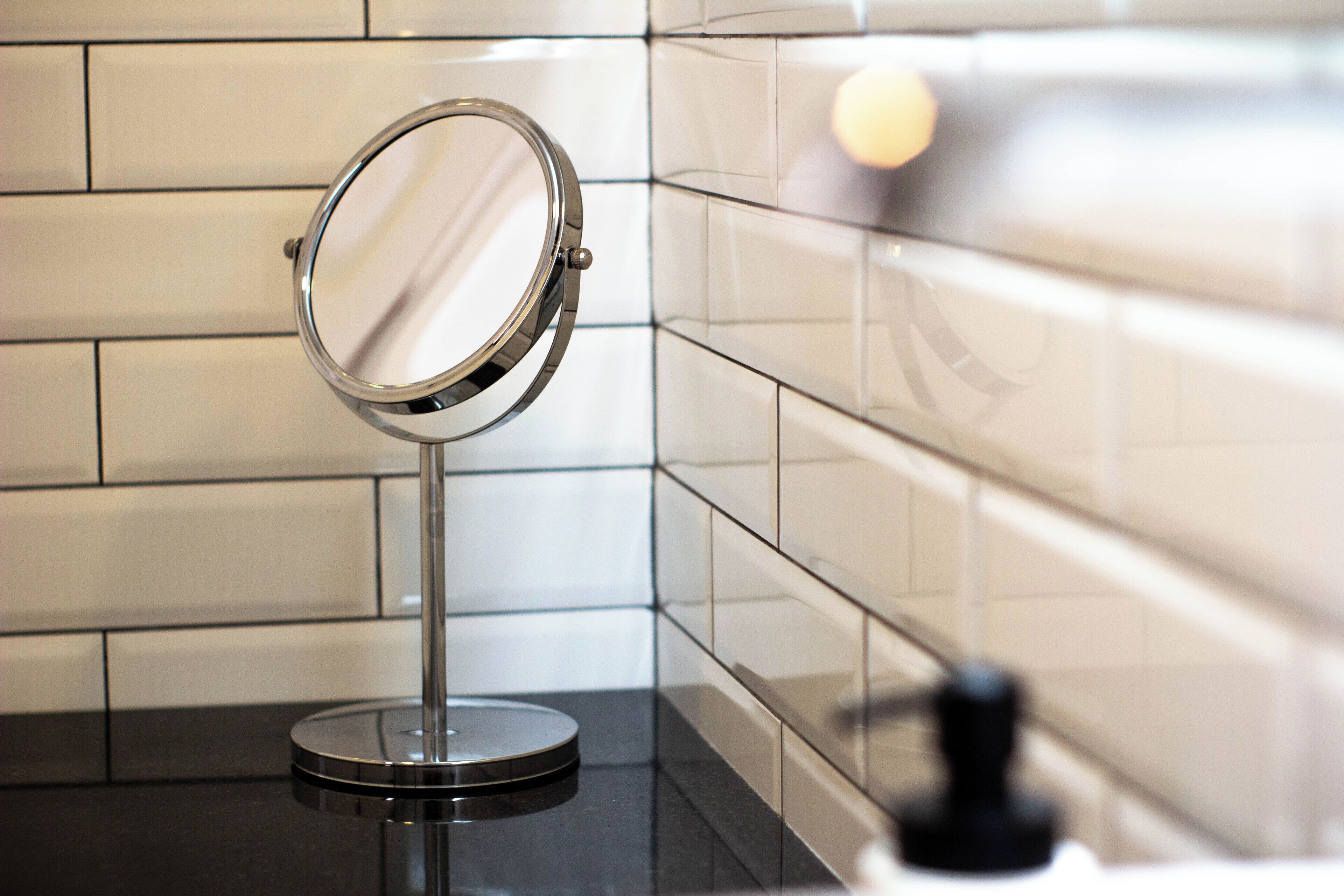 A bathroom with a round mirror on a stand, tiled walls, and a black countertop.