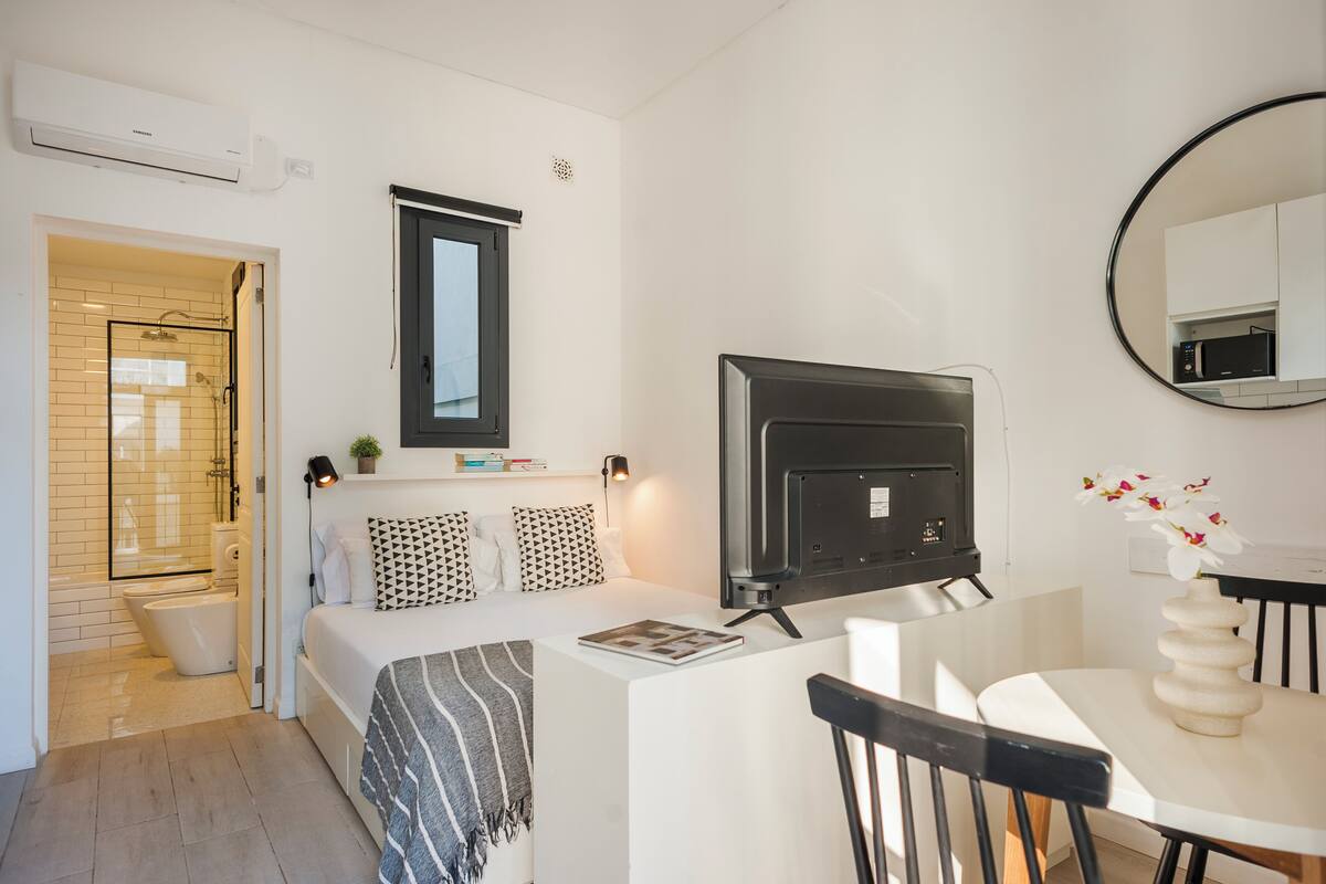A modern bedroom with a TV, a bedside table, a chair, a small table, and a mirror.