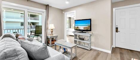Un salon avec un canapé gris, un téléviseur posé sur un meuble en bois et un balcon doté de balustrades blanches.