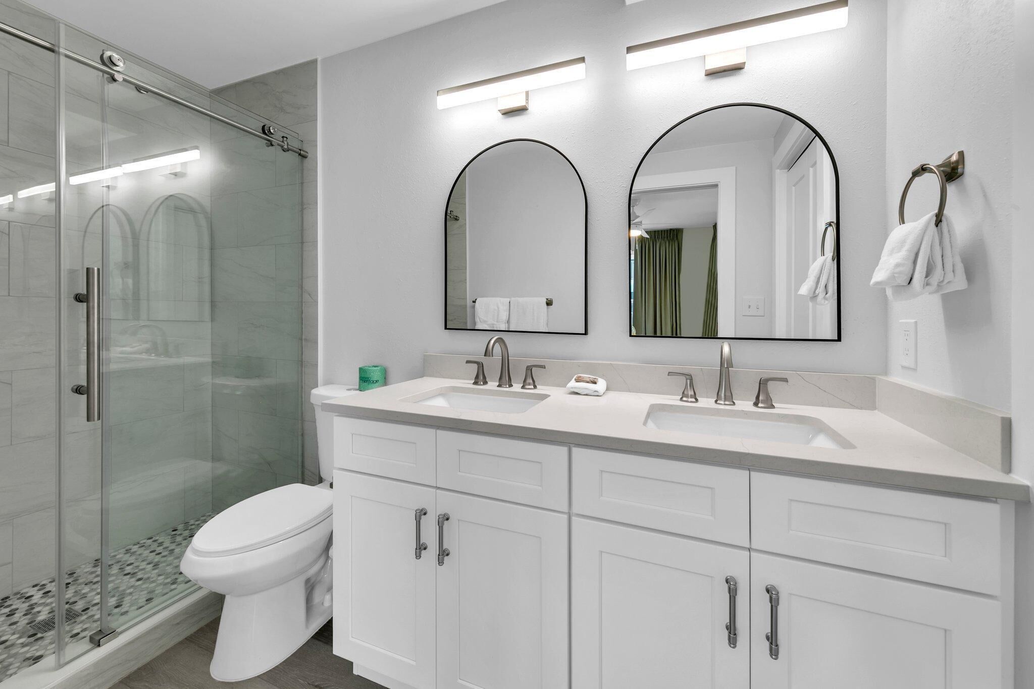 A modern bathroom with a glass shower enclosure, a white toilet, and a vanity with two sinks and a large mirror.