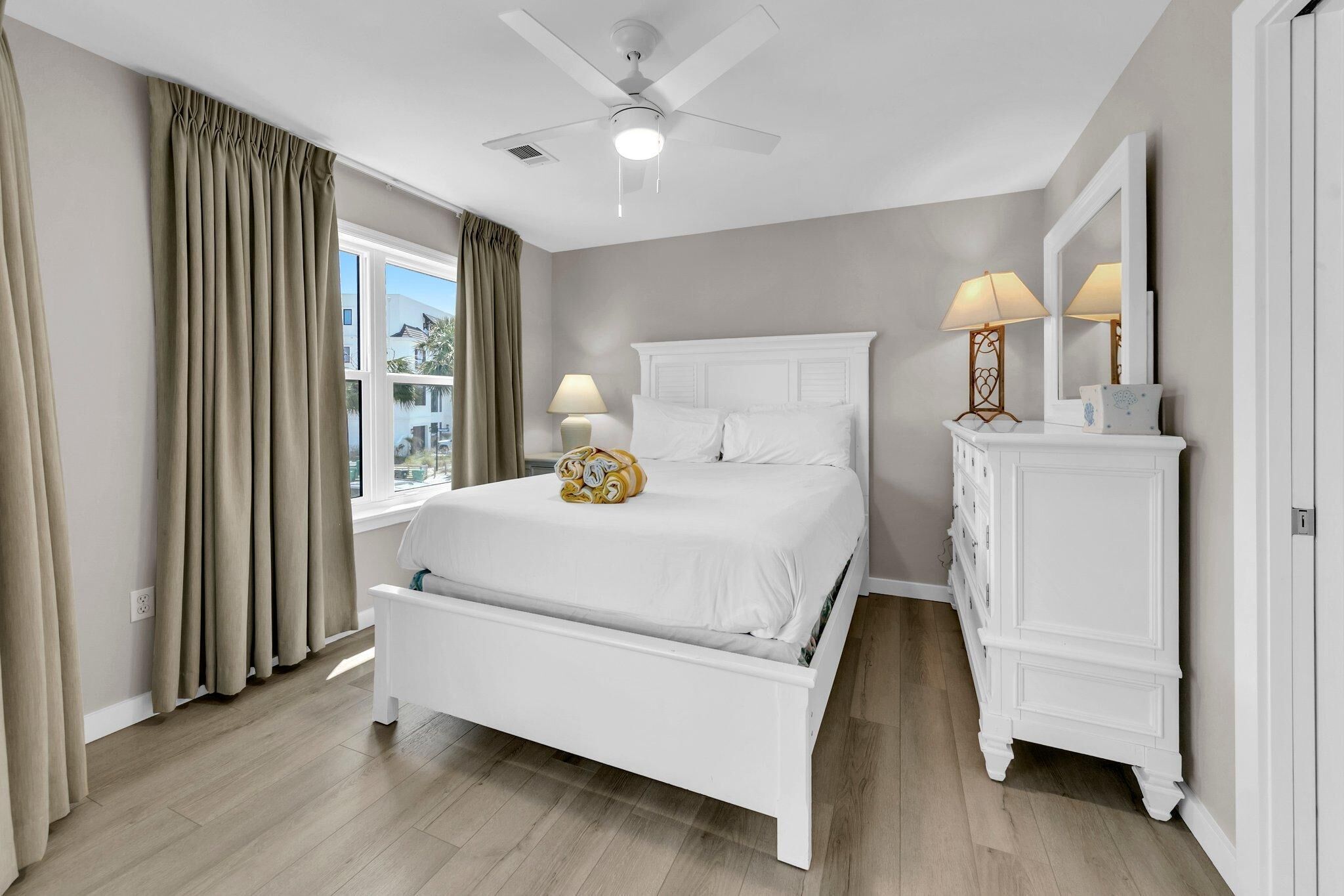 A bedroom with a white bed, a ceiling fan, a dresser, and a window with curtains.