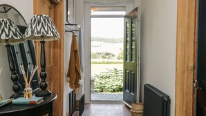 Ein Flur mit Holzboden, einem Heizkörper, einem Tisch mit Lampe und Blick auf eine grüne Landschaft durch eine offene Tür.