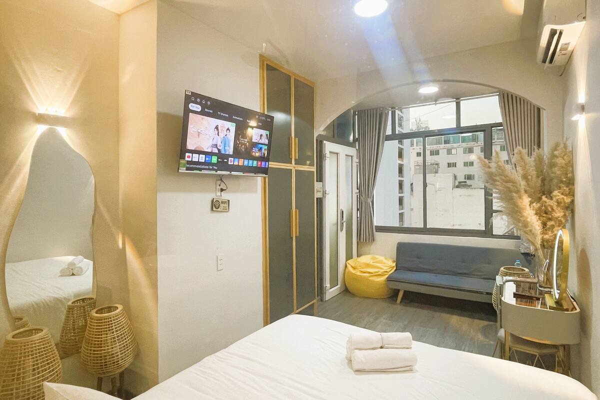 A hotel room with a bed, a TV mounted on the wall, a window with curtains, a sofa, and a vase with pampas grass.