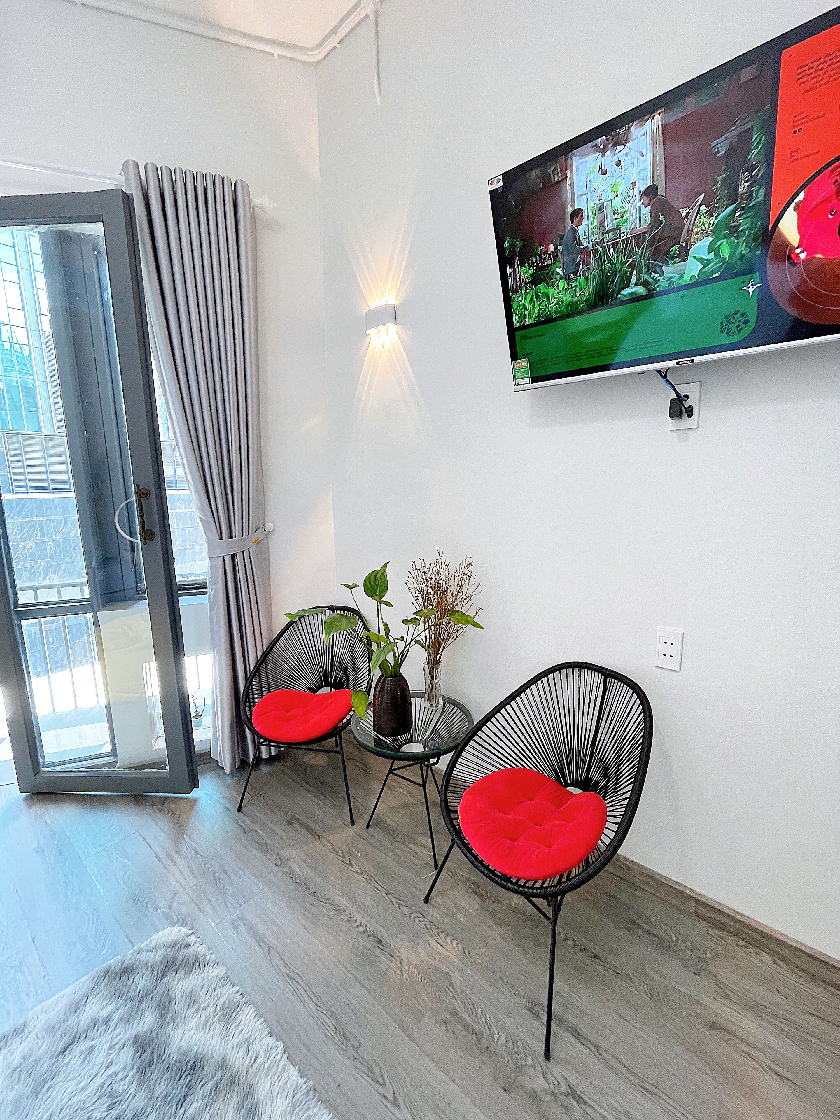 A room with two black wireframe chairs with red cushions, a small round table, a potted plant, and a flat-screen TV mounted on the wall.