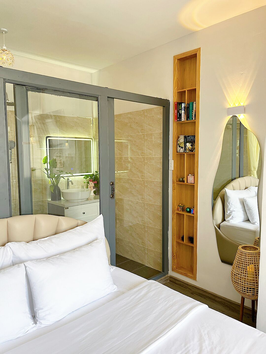 Una habitación de hotel con cama, baño con mampara de vidrio, estante de madera con artículos de aseo, espejo redondo y una silla de mimbre.
