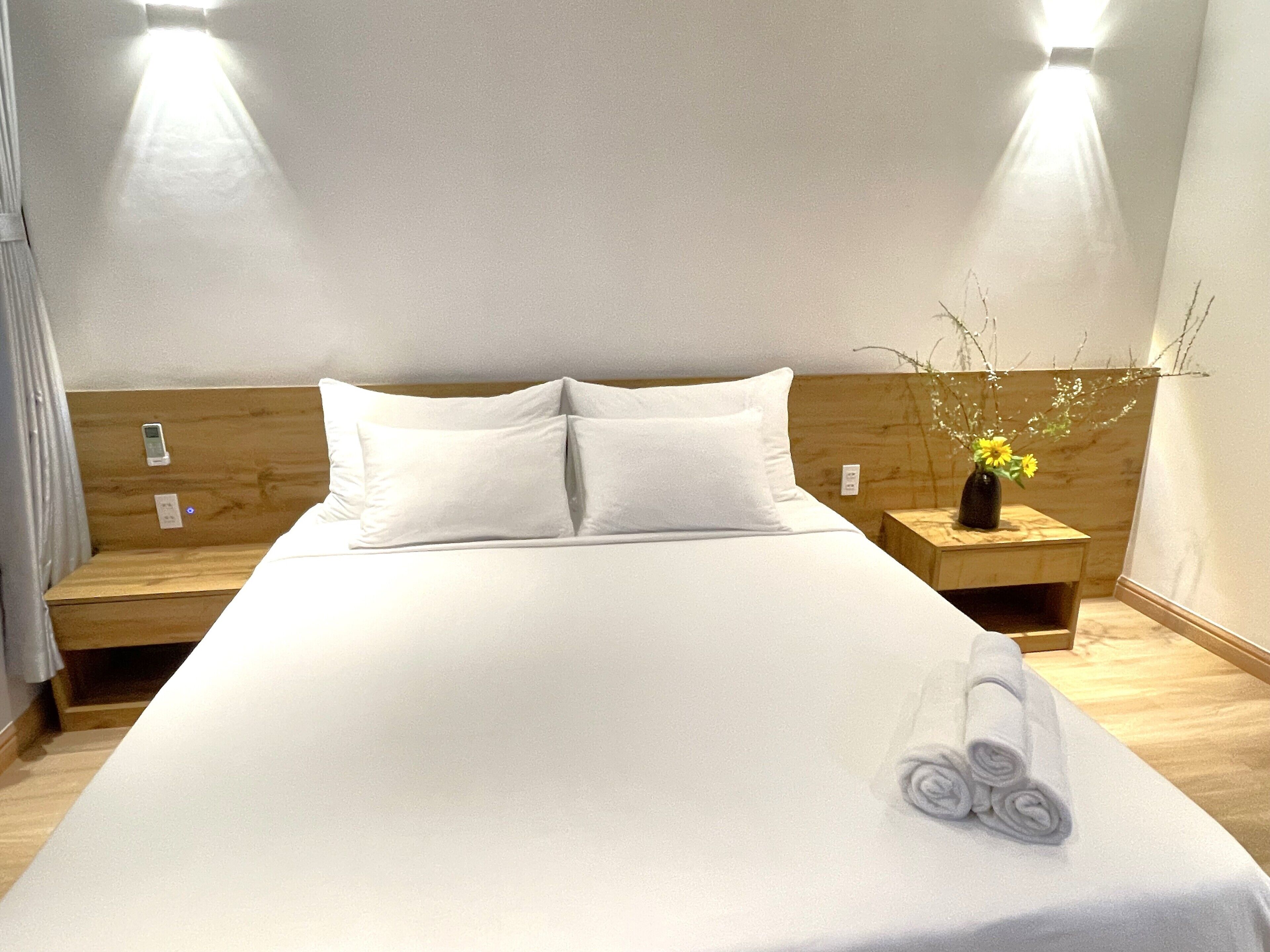A neatly made bed with white linens, two pillows, and a rolled towel on the side. There is a wooden headboard and a nightstand with a vase of flowers.
