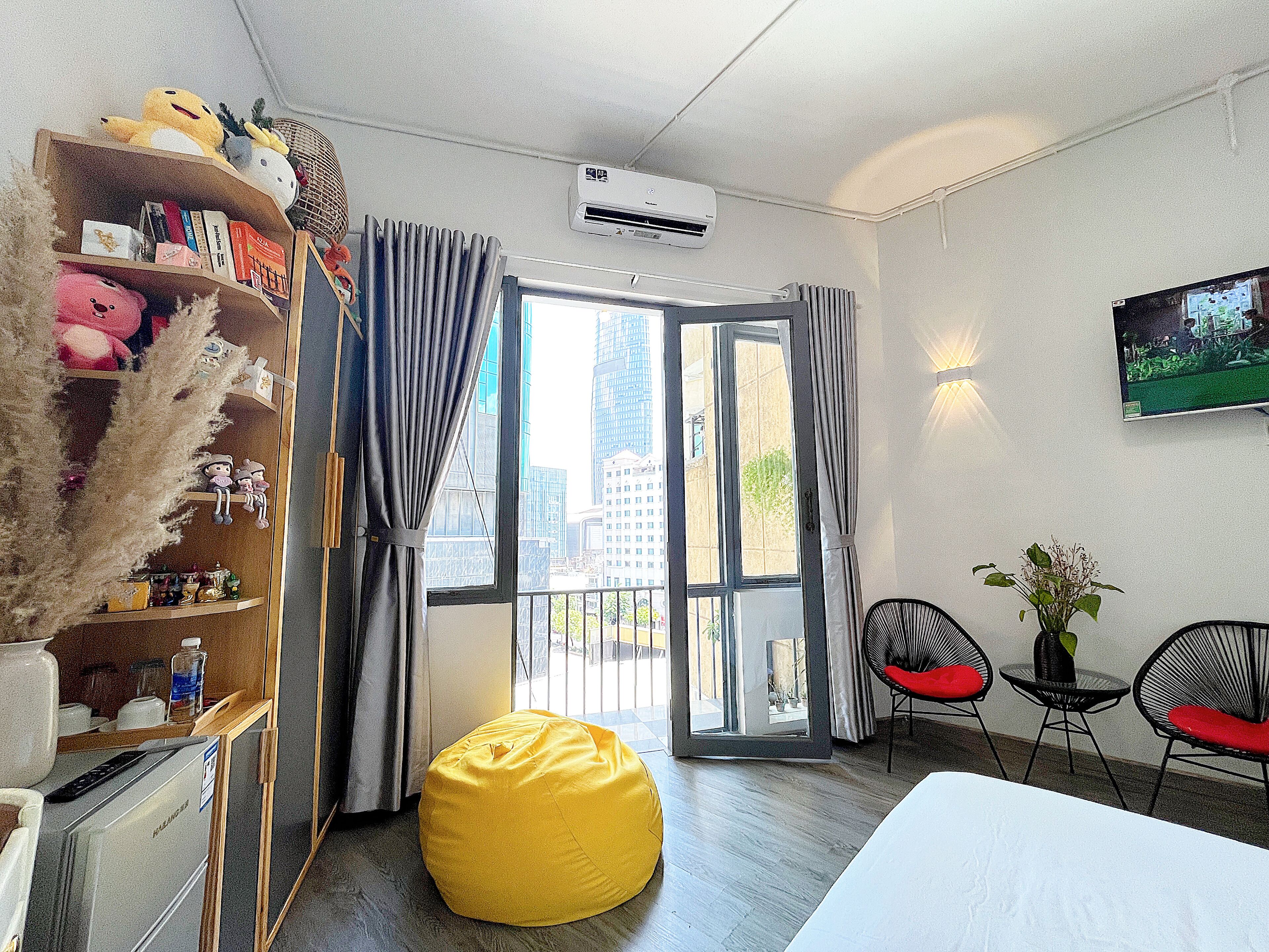 A modern hotel room with a large window, a yellow bean bag, a bookshelf with plush toys, and a TV on the wall.