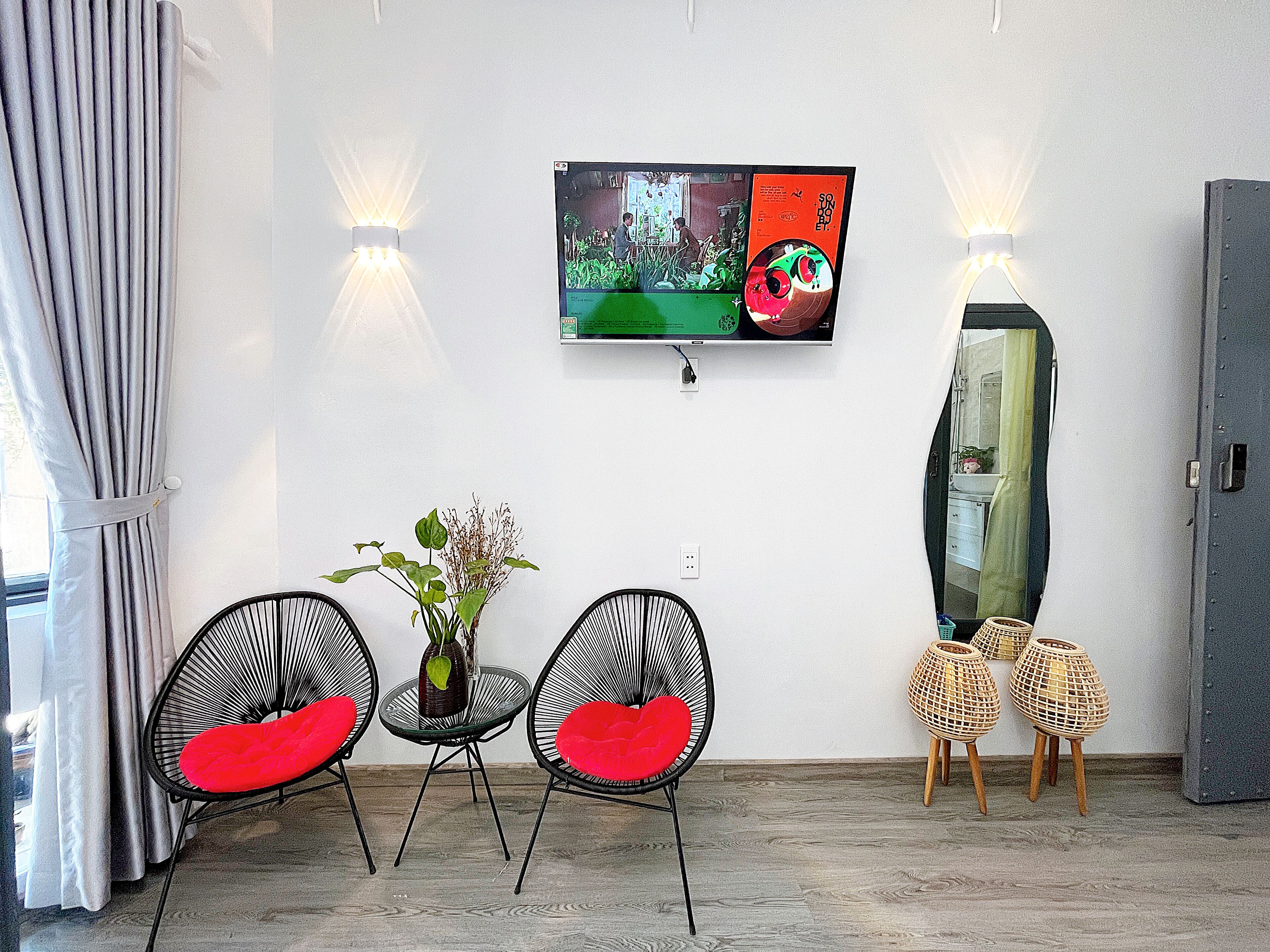 A modern living room with a flat-screen TV mounted on the wall, two black chairs with red cushions, a small round table with a vase, and a tall cabinet.