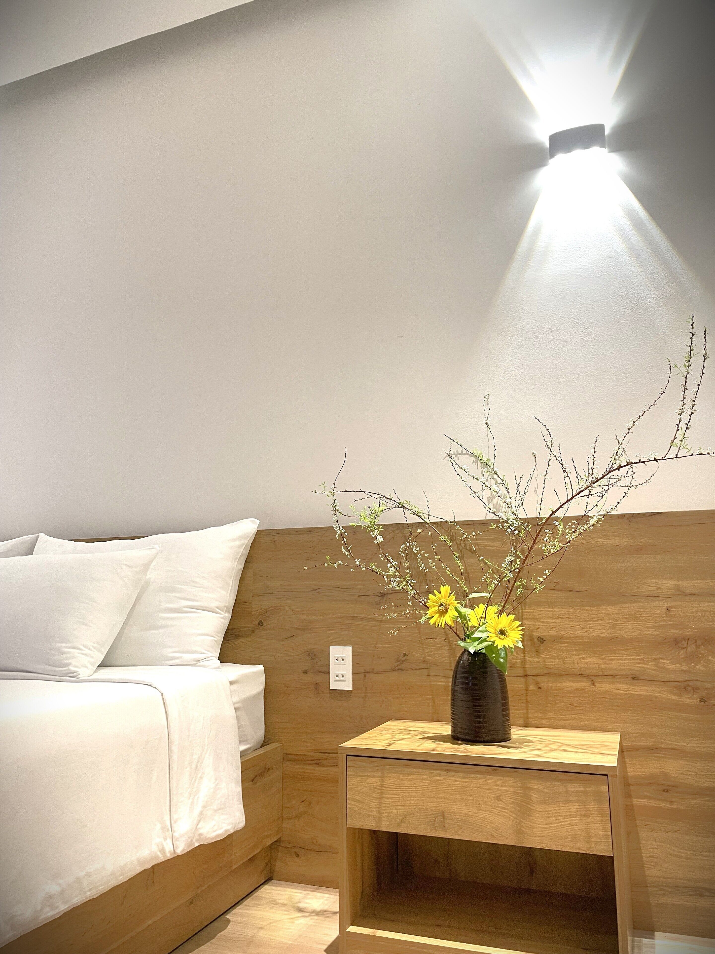 Une chambre à coucher avec une tête de lit en bois, une table de chevet ornée d’un vase de fleurs et un luminaire fixé au mur.