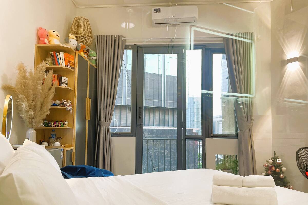 A hotel room with a bed, a bookshelf, a window with curtains, and a small Christmas tree.