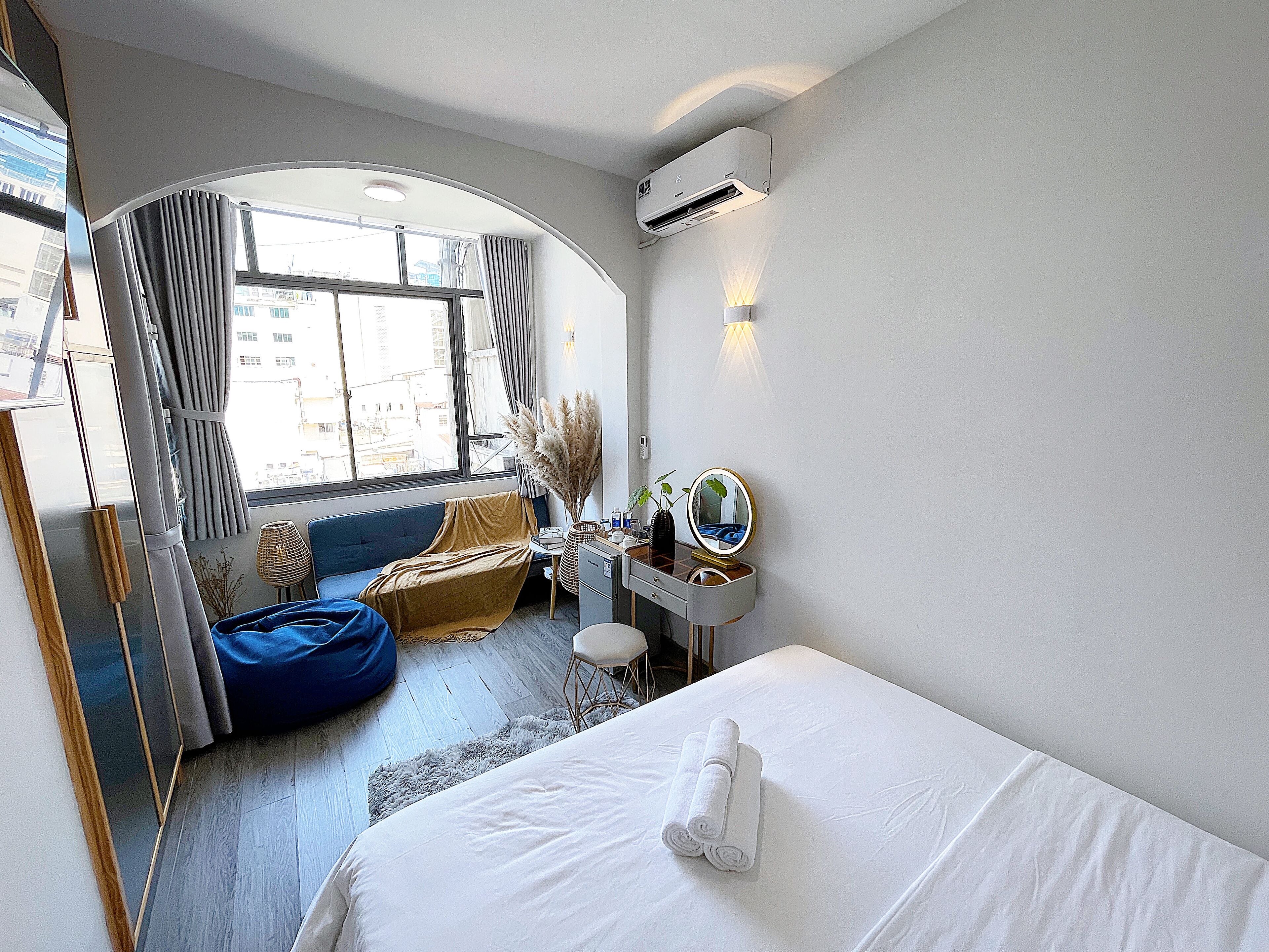 A hotel room with a bed, a desk with a mirror, a chair, a sofa, and a large window with curtains.