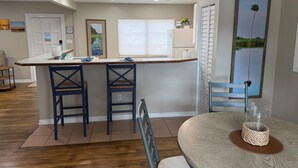 Una cocina con barra, dos taburetes, una mesa redonda con un centro de mesa tejido y un ventanal con persianas.
