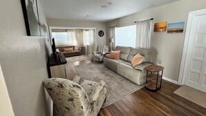 Un salón amplio con un sofá grande, un sillón estampado y una mesa de centro. Hay un comedor con una mesa y sillas, y una zona de cocina con un refrigerador y una estufa.
