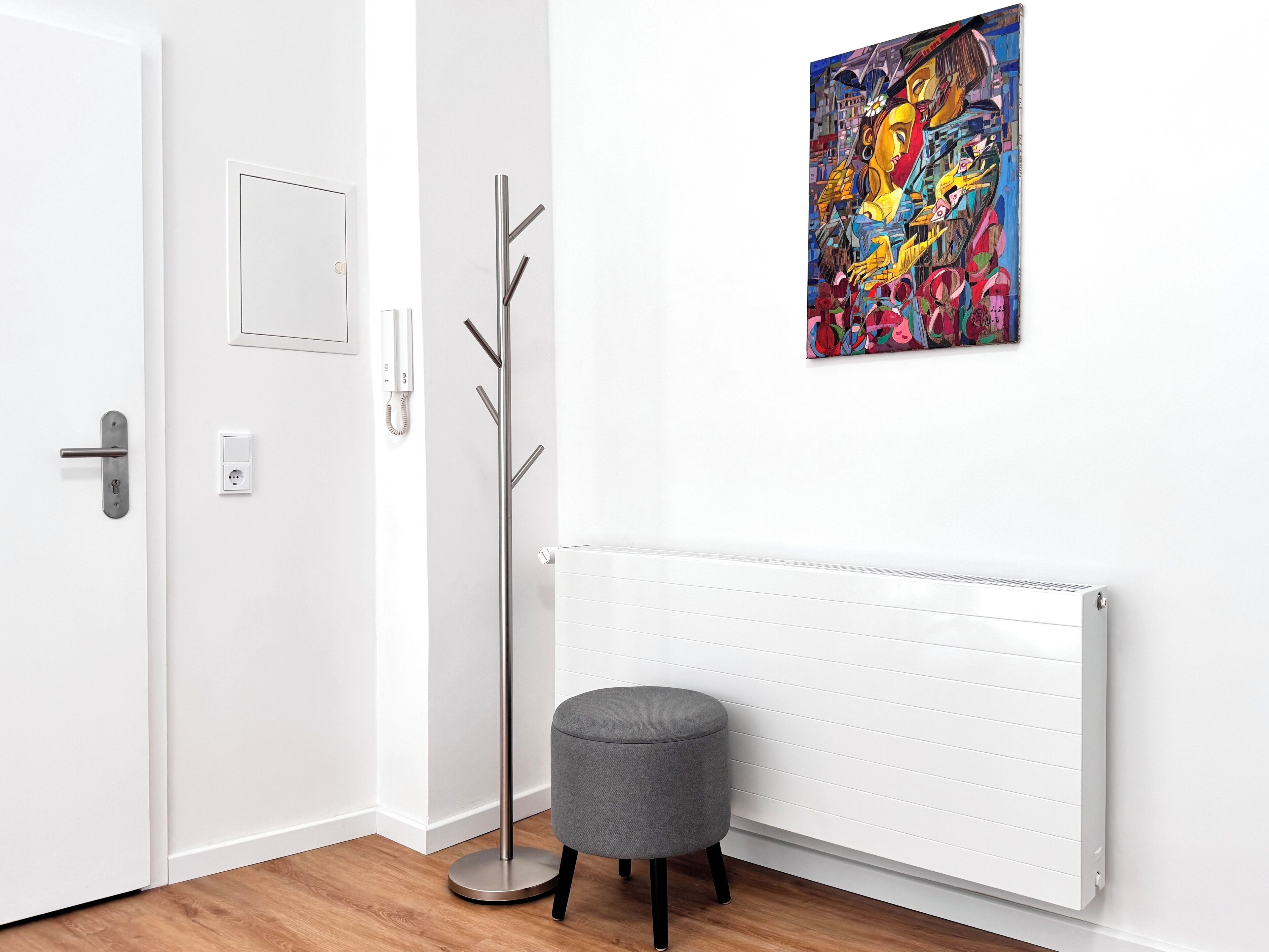 A modern room with a white door, a coat rack, a grey ottoman, a radiator, and a colorful abstract painting on the wall.