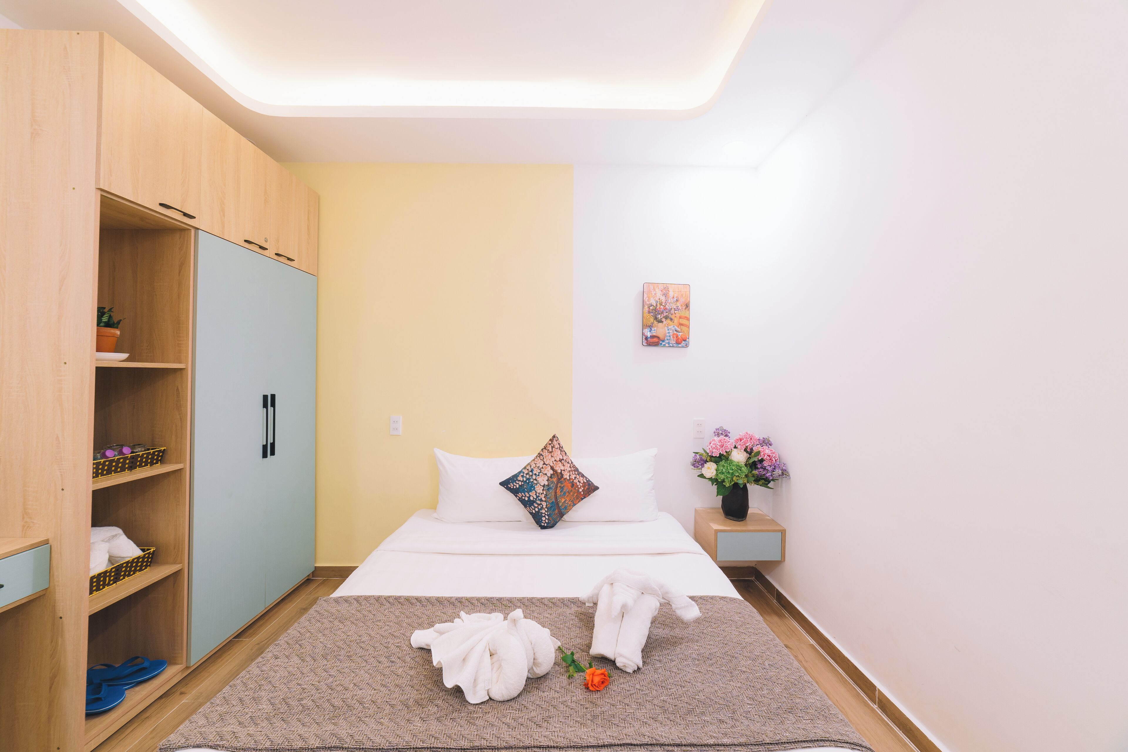 Una habitación de hotel con cama, un armario, una mesita con un florero y un cuadro en la pared.