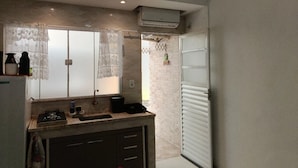 A compact kitchen with a gas stove, sink, and a small countertop. There is a window with lace curtains, an air conditioning unit above, and a sliding door leading to another room. - Suítes e Kits da Natasha (Arraial do Cabo)