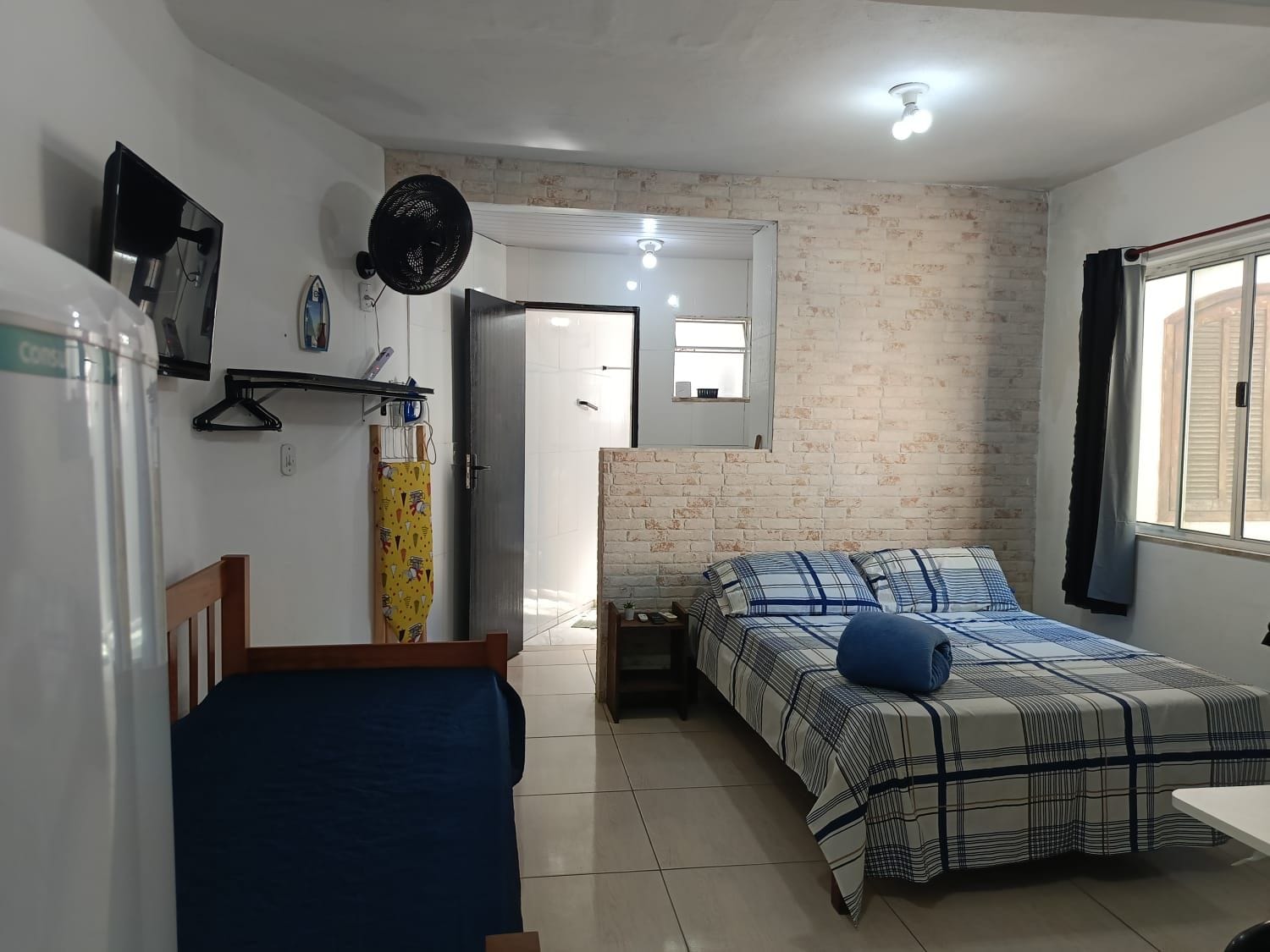 Un dormitorio con cama, mesita de noche, ventilador de techo y ventana con cortinas.