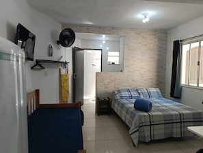 A bedroom with a bed, a bedside table, a hanging fan, and a window with curtains. - Suítes e Kits da Natasha (Arraial do Cabo)