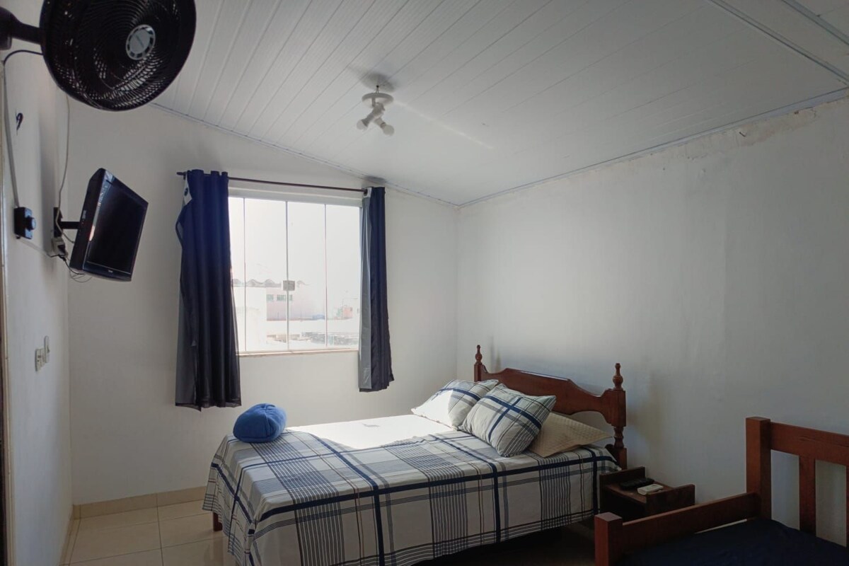 A bedroom with a bed, a television mounted on the wall, a ceiling fan, and a window with curtains.