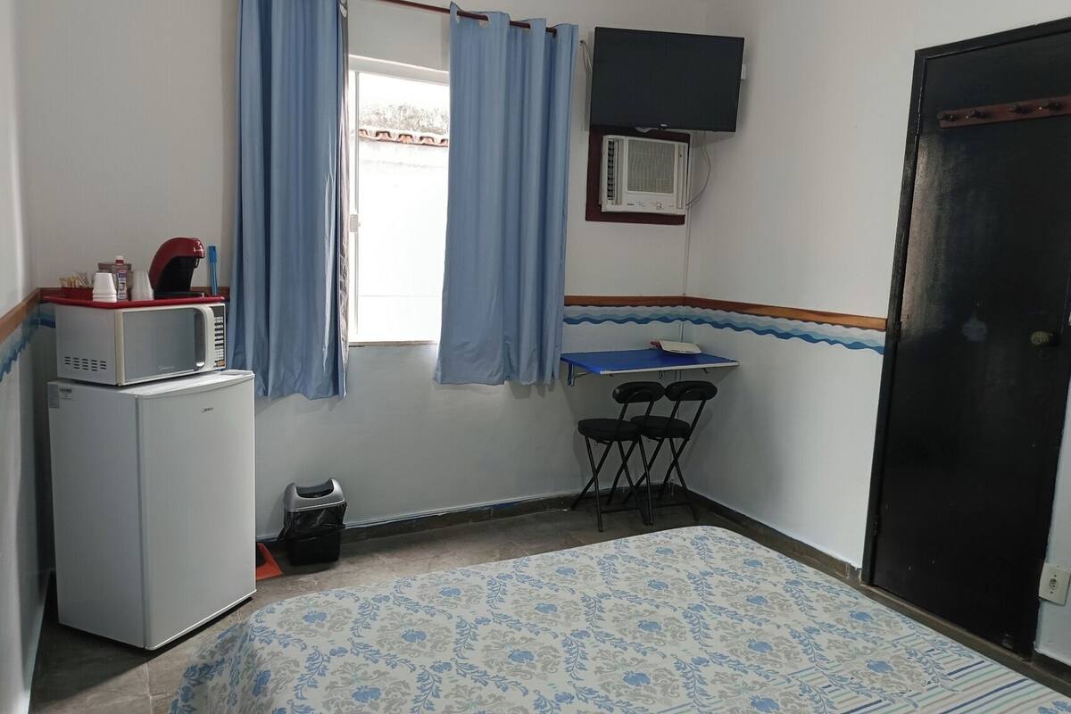 Una habitación pequeña y limpia con una cama, un escritorio, una silla, un microondas y un cubo de basura.