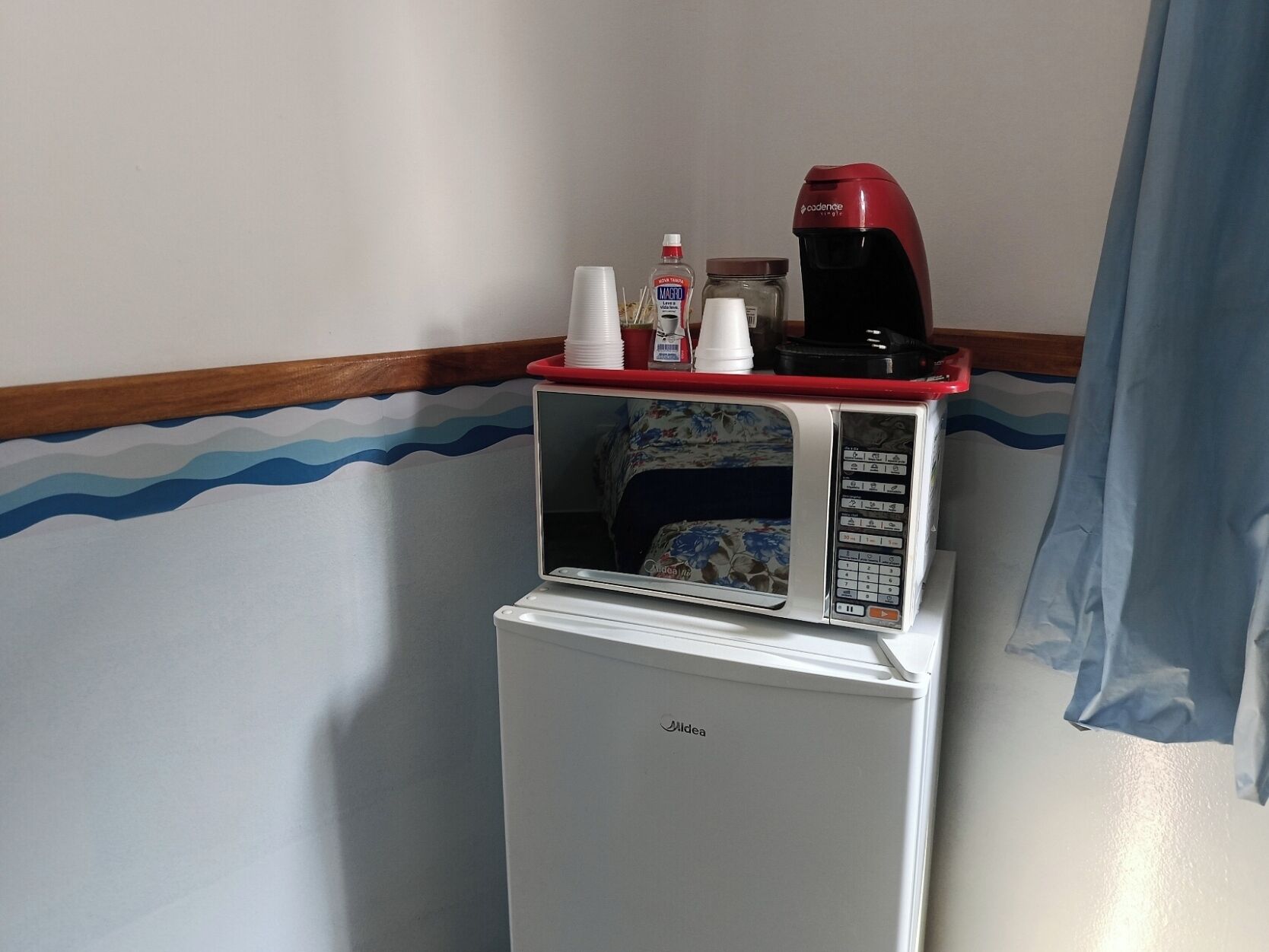 A minibar with a microwave and a coffee maker, a bottle of hand sanitizer, and a stack of white cups.