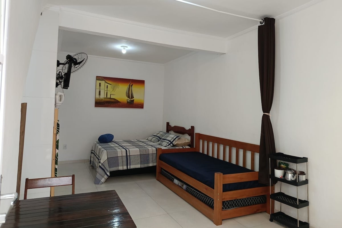 A small, compact room with a bed, a chair, a table, and a shelf with kitchen items.