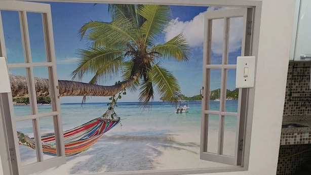 A room with a large window revealing a beach scene with a hammock, palm trees, and a boat. - Suítes e Kits da Natasha (Arraial do Cabo)