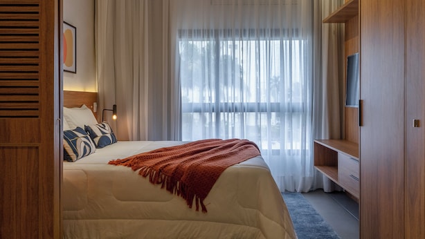Ein Schlafzimmer mit einem Bett, einem Fernseher, einem hölzernen Kleiderschrank und einem großen Fenster mit transparenten Vorhängen.
