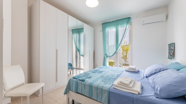 A bedroom with a bed, a chair, a wardrobe, and a window with curtains. - Villetta Soave - Santo Isidoro (Santo isidoro)