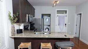 Une cuisine avec des meubles de couleur sombre, un plan de travail en granit, un lave-linge et deux tabourets de bar.