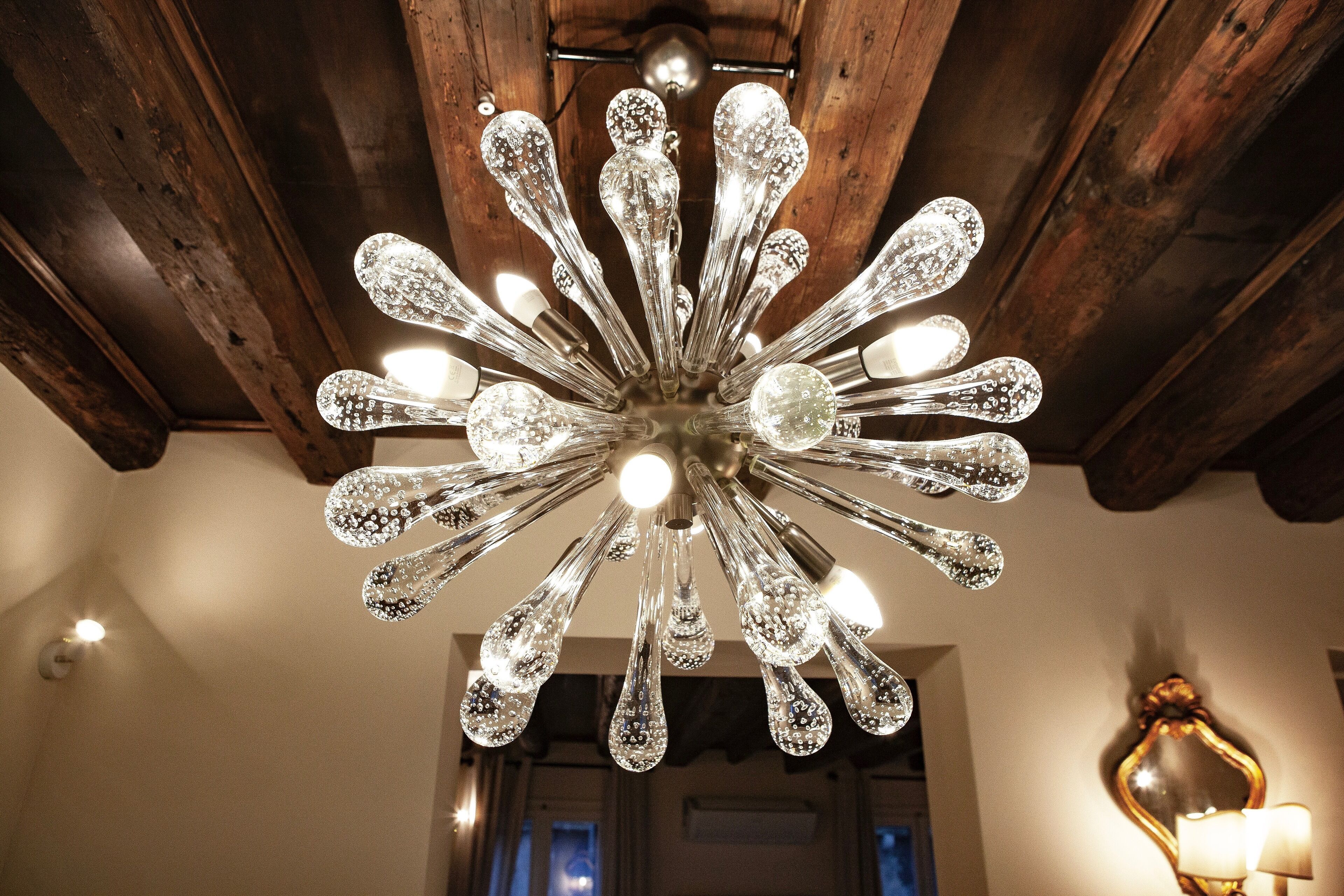 A chandelier with multiple arms and crystal-like bulbs in a room with wooden beams and a mirror.