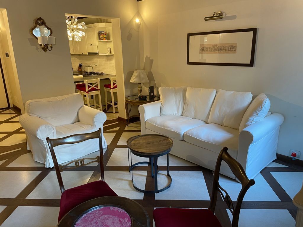 A living room with a white sofa, two armchairs, a small round table, and a kitchen area in the background.