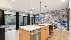 A modern kitchen with a central island, stainless steel dishwasher, pendant lights, and a cityscape mural on the wall.