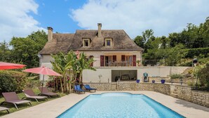 Een zwembad met ligbedden en parasols, een huis met balkon, en een stenen muur.