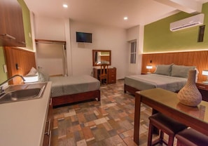 Una habitación de hotel compacta con dos camas individuales, una kitchenette pequeña, un espejo y un jarrón decorativo.