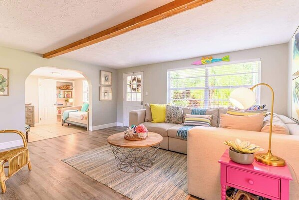 A cozy living room with a sofa, a coffee table, and a pink side table. There is a window with blinds and a door leading to another room.
